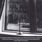 Fotografie alb-negru realizată în 1931 de Rachel Posner, soția rabinului Akiva Posner. Imaginea surprinde un menorah de Hanukkah cu lumânările aprinse, așezat pe un pervaz de fereastră. În fundal, prin fereastră, se vede un steag nazist arborat pe o clădire. Fotografia simbolizează rezistența spirituală și identitatea evreiască în fața opresiunii naziste. Sursa imaginii: Yad Vashem.